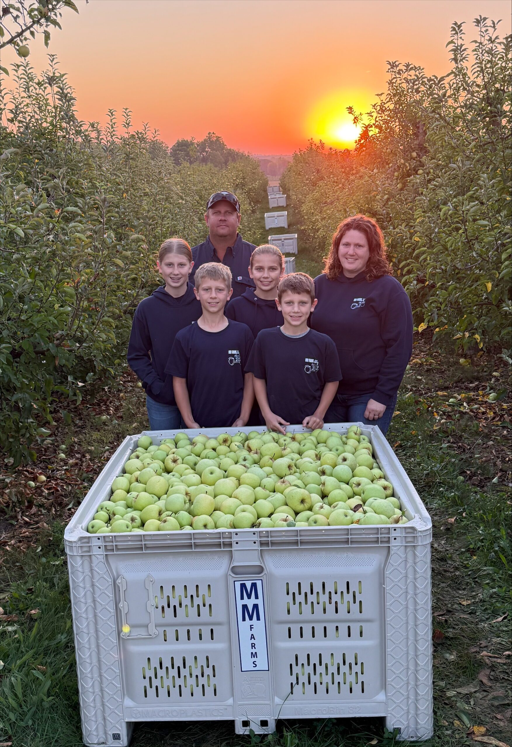 Photo of Kyle Weber and Melissa Morlock with their children in apple orchard in front of apple harvesting bin.