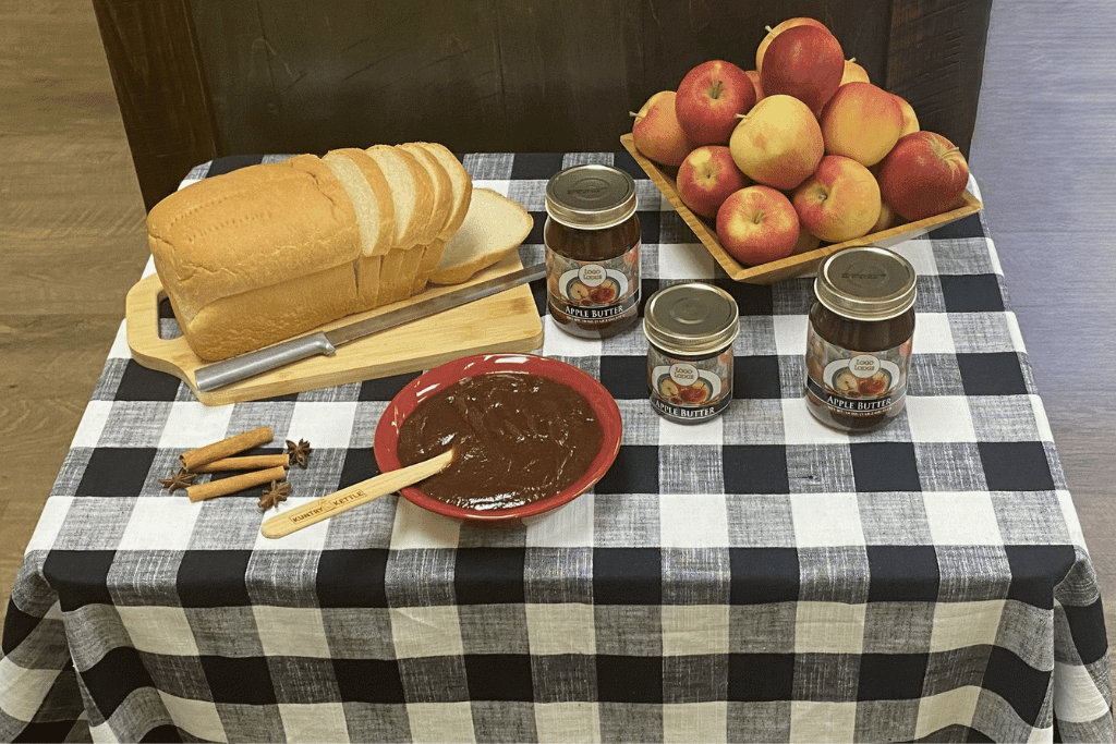 Image depicts a sampling table with jars of jam, apples and a loaf of bread